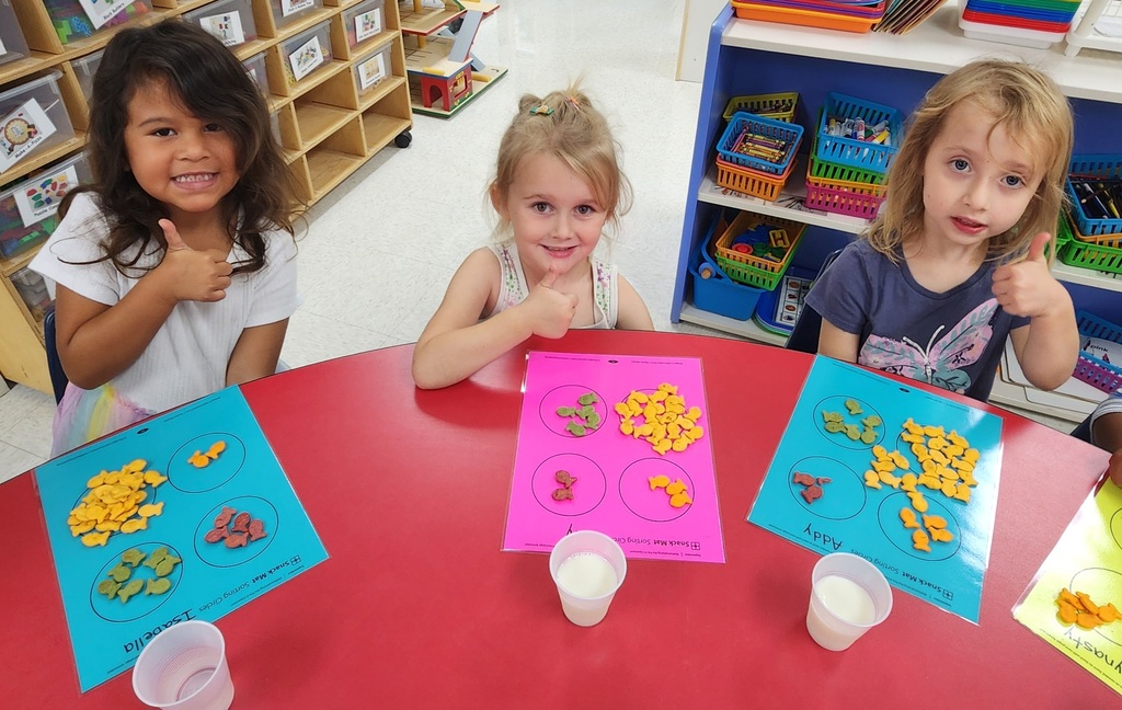Three young learners sitting at a table, giving the thumbs up with goldfish crackers sorted in front of them.