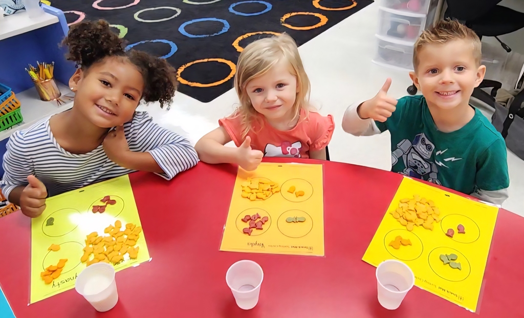 Three young learners sitting at a table, giving the thumbs up with goldfish crackers sorted in front of them.