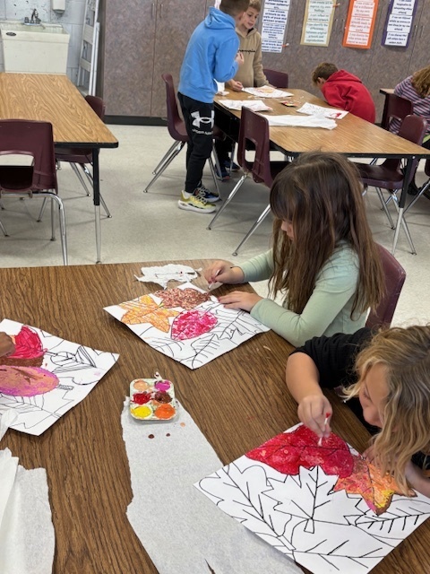 Elementary students working on art work at a table with paint trays in front of them.