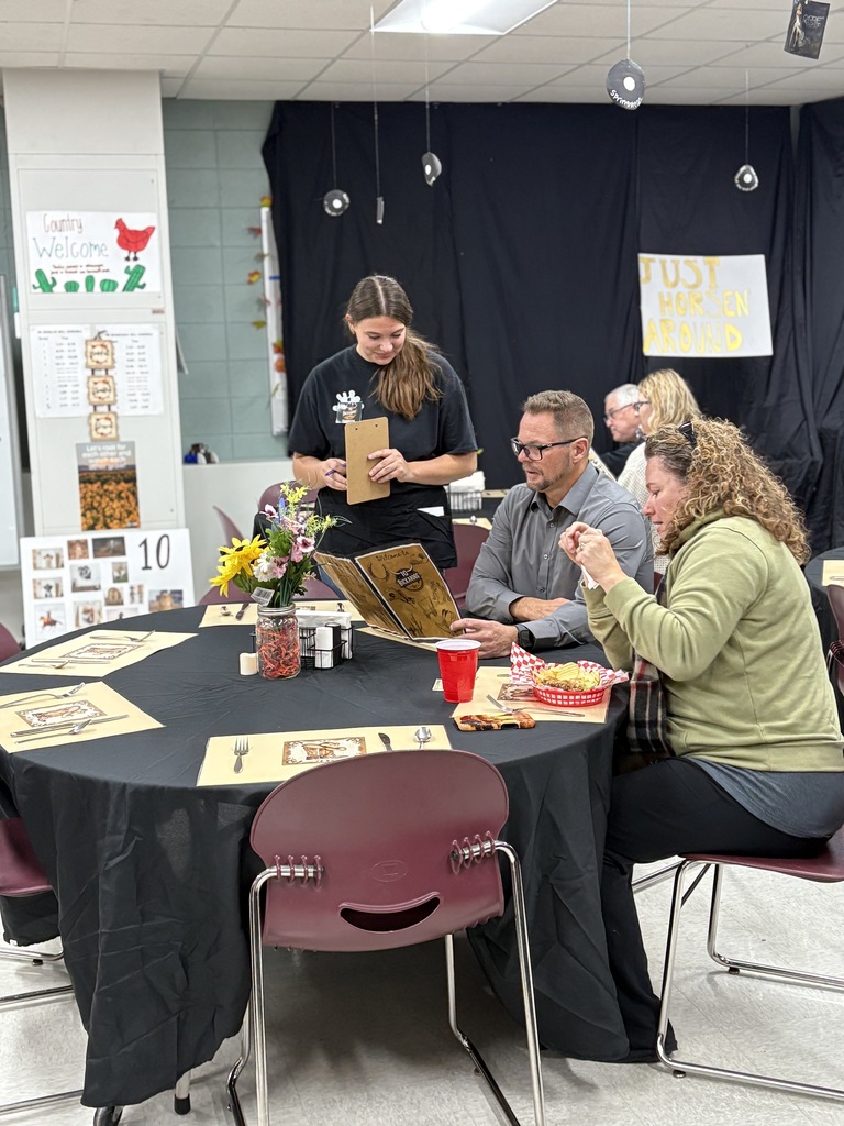 Two adults dining at a table with a high school server talking with them. 