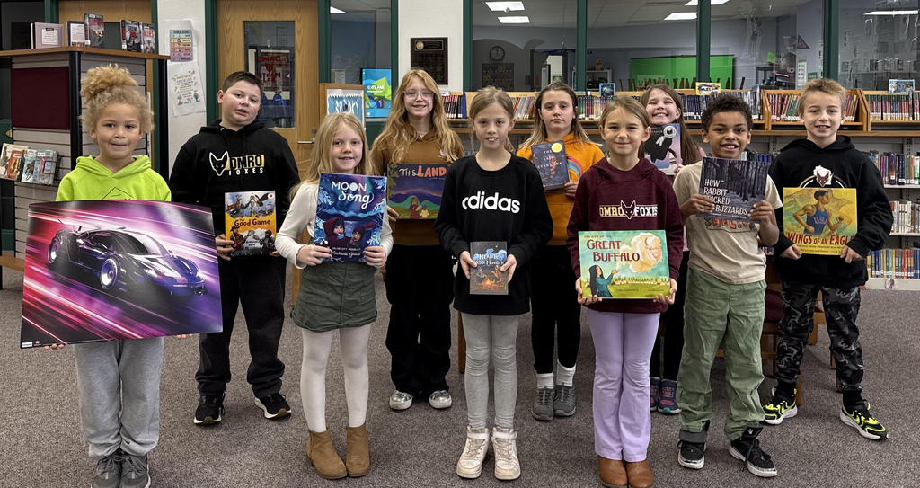 A group of elementary students standing in a media center, holding a book or poster.