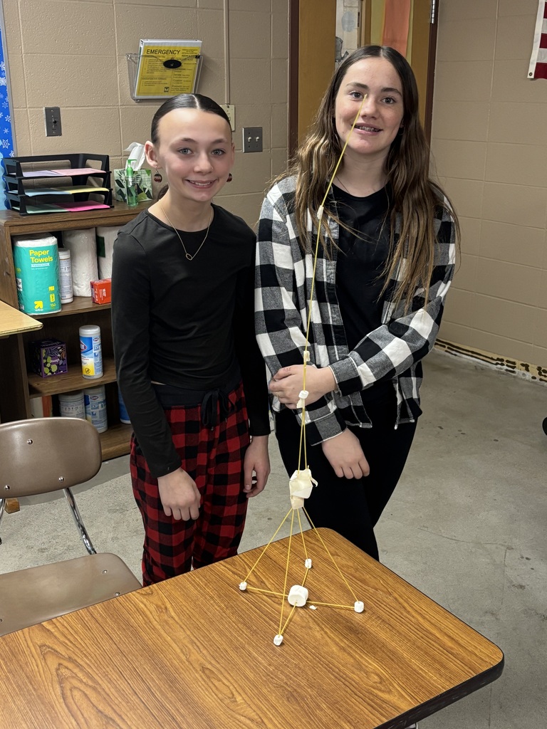 Two middle school girls standing by their marshmallow and spaghetti noodle structure that is on a table. 