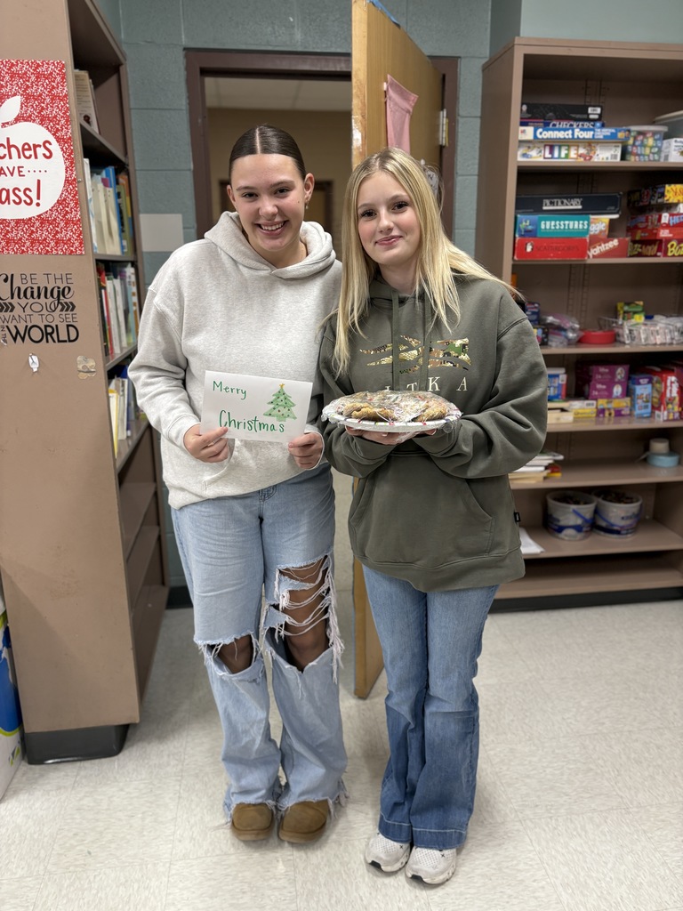 2 high school students standing and holding a plate of cookies with a sign that says, "Merry Christmas."