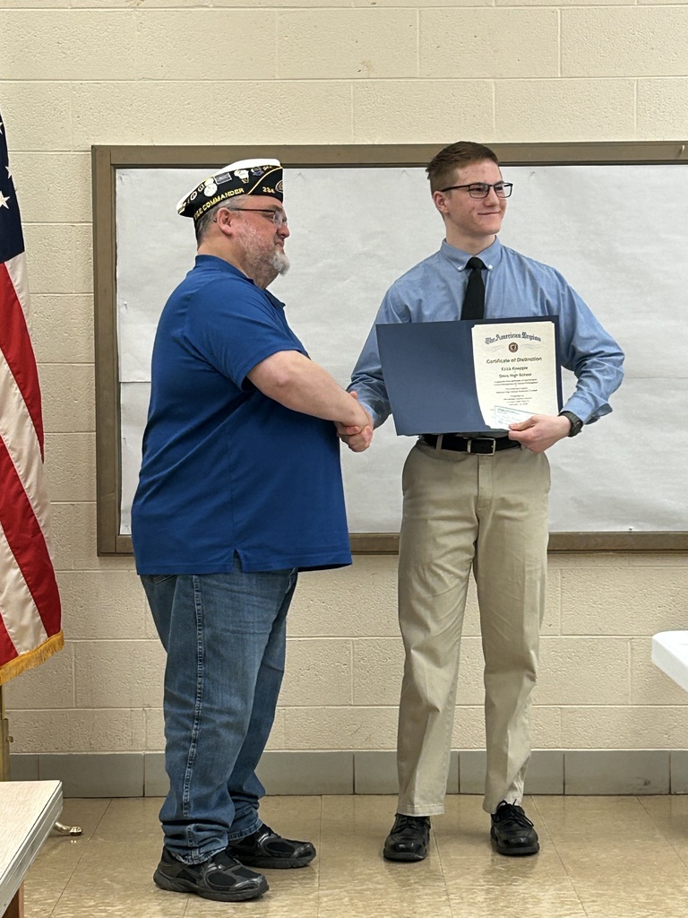 High school student dressed in nice clothing shaking the hand of an American Legion member near a United States of America flag.