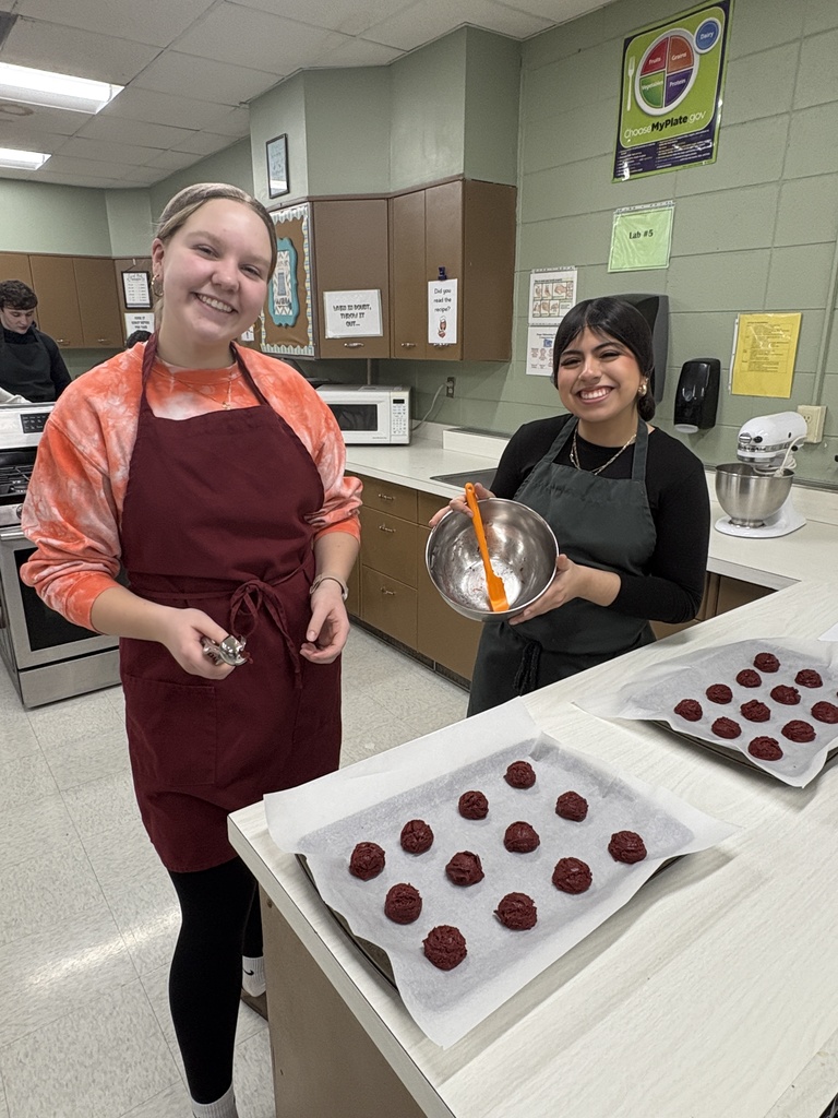 Two high school students in a school kitchen with cookie dough balls on trays in front of them.