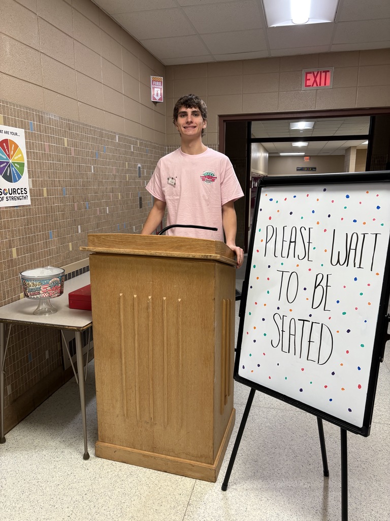 High school student standing behind a podium near a sign that says "Please Wait to be Seated".
