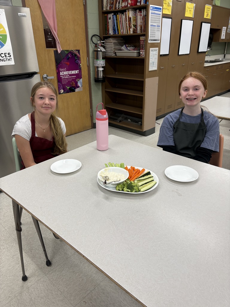 Two middle school students sitting at a table with a plate of vegetables and dip in front of them. 