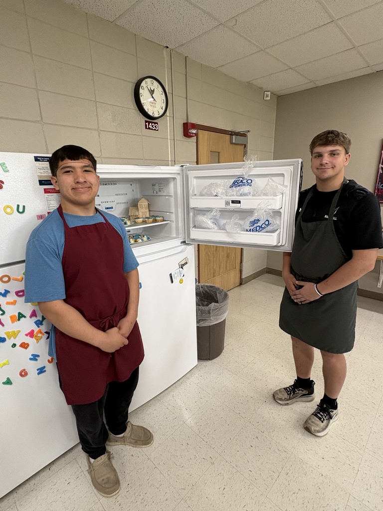 Two high school students standing near a fridge with a freezer door open that has cupcakes in it.
