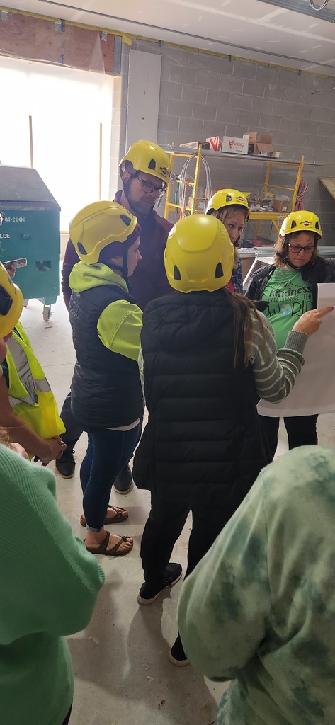 A small group of adults standing in a circle, wearing hard hats and looking at plans. 