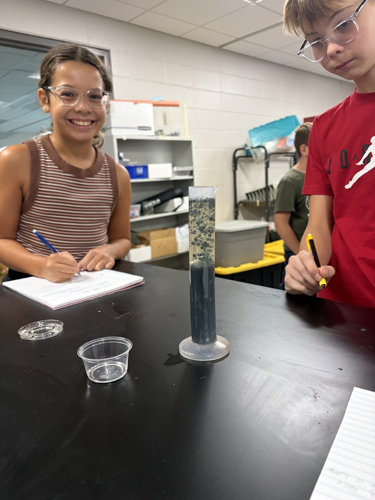 Two middle school students at a lab table with notebooks and a test tube of lava lamp material in front of them.