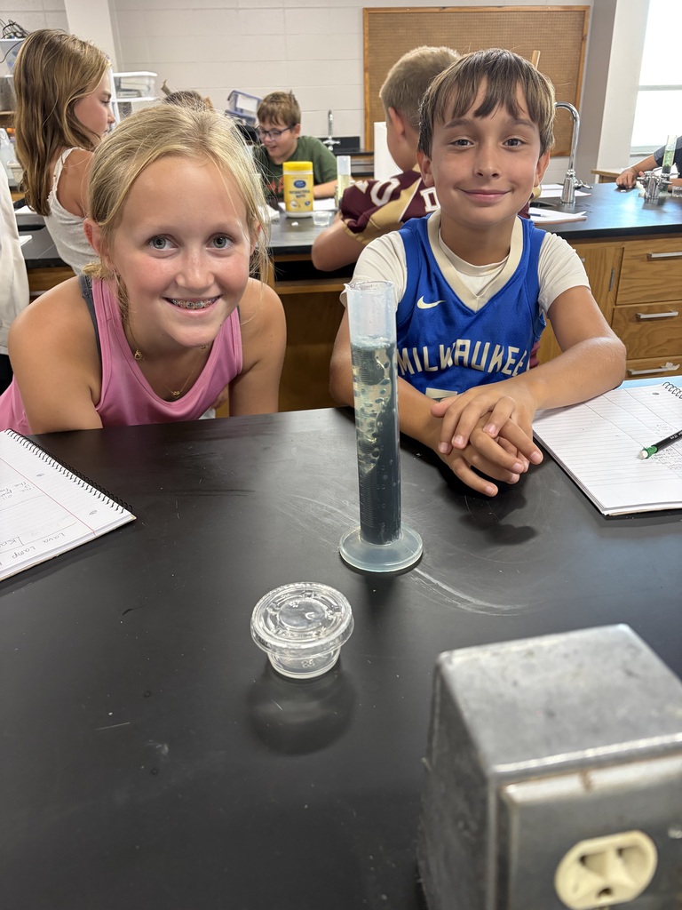 Two middle school students at a lab table with notebooks and a test tube of lava lamp material in front of them.