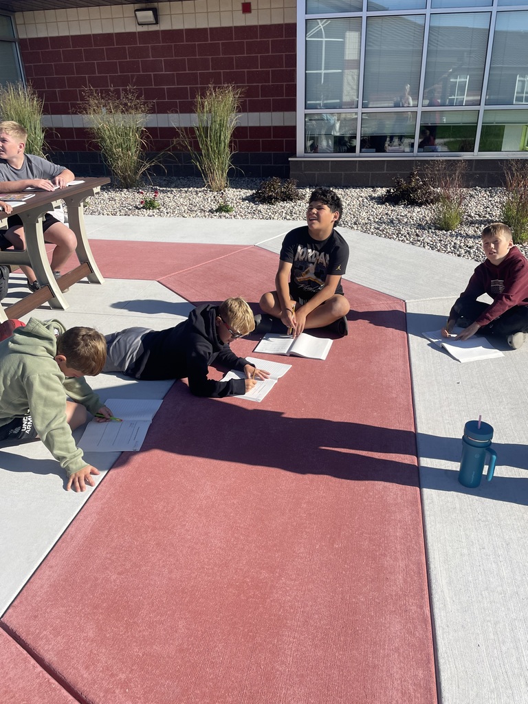Elementary students sitting at tables and laying on the ground while utilizing an outdoor learning space.
