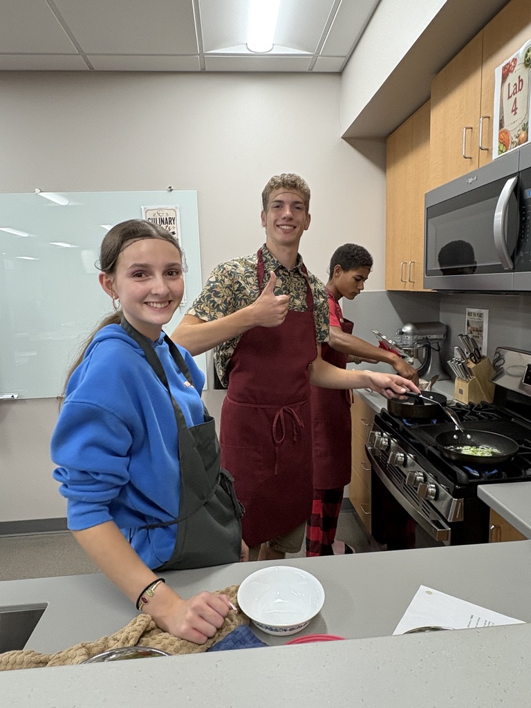 Three high school students standing in a Food Lab kitchen preparing food.