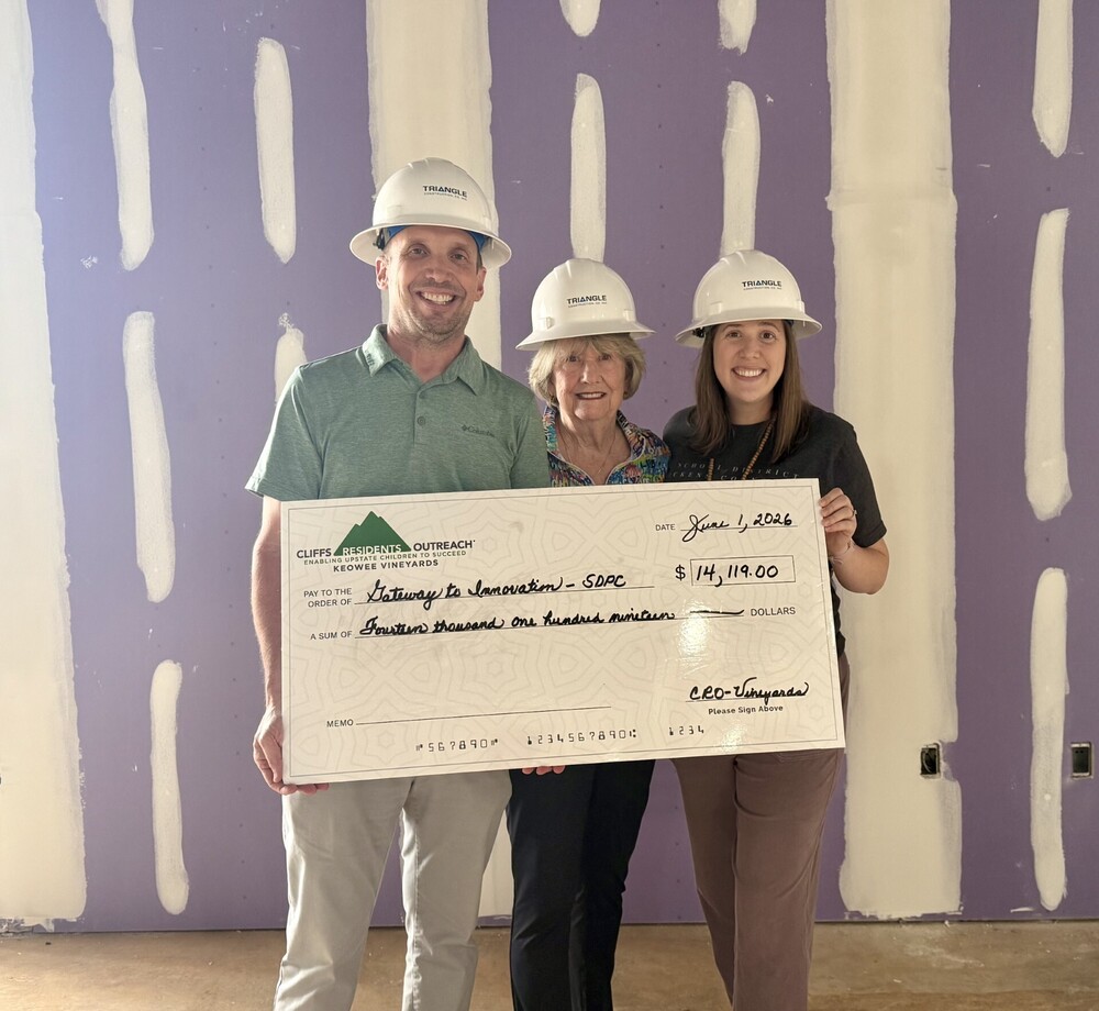 Tim Johnson, Char Burgner, and Katie Britton are holding a large check from the Keowee Vineyards Cliffs Residents Outreach. All are wearing hardhats and standing in front of an unfinished wall .i