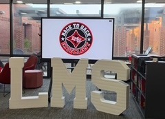 A celebration setup at Liberty Middle School featuring a red carpet flanked by large black, red, and white balloon arches, leading to giant lit-up "LMS" marquee letters and a screen displaying a "Back-to-Back" logo.