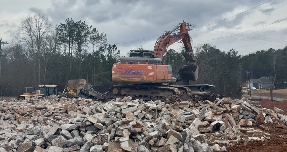 G2i Site- Grinding Concrete and Asphalt. Pile of asphalt with a trackhoe on top loading concrete into grinder
