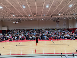 **ADA-compliant alt text:**  “Large group of students and staff seated in the bleachers of a school gymnasium during a morning meeting, with a basketball court and stage area visible in the foreground.”