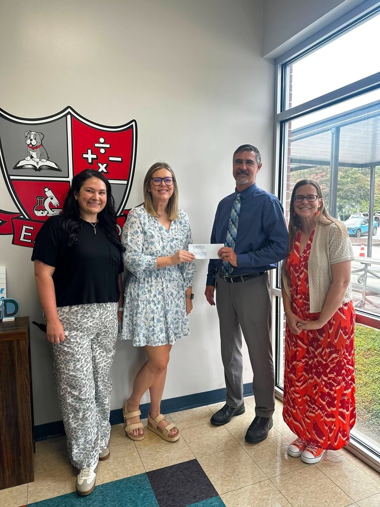 Three East End Elementary staff members and United Way Member stand together near a large school logo, smiling as one staff member presents another with a check. The photo highlights support for a school program or initiative.