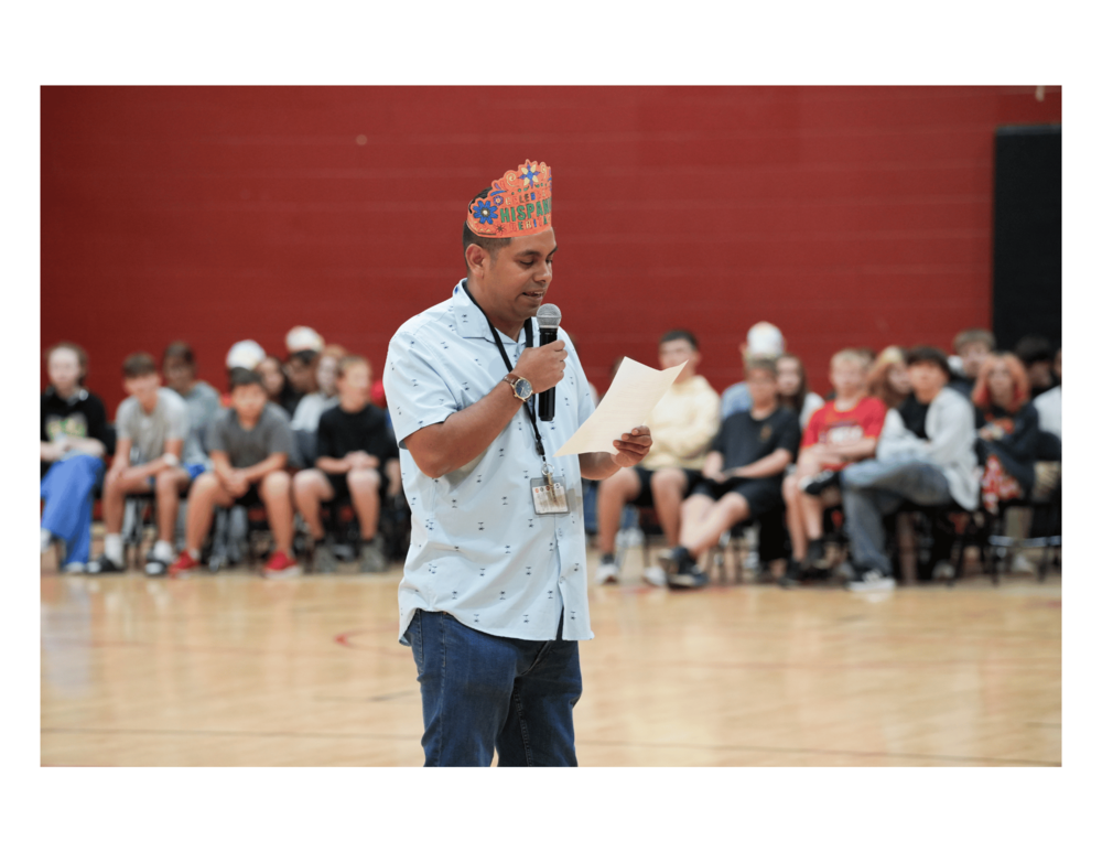 A Liberty Middle School Spanish teacher stands in a gymnasium holding a microphone and reading from a paper during the school’s Hispanic Heritage Celebration. He wears a colorful Hispanic Heritage crown, while students and staff sit in the background.