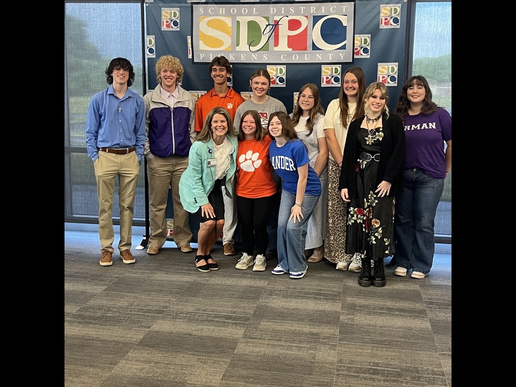 Group photo of PHS seniors who plan to major in education, posing with Dr. Lucas, Teacher Cadet sponsor, in front of a School District of Pickens County (SDPC) backdrop. Dr. Lucas, wearing a teal blazer, kneels in front beside two students, while the remaining students stand behind. The group is smiling and dressed in a mix of casual and school-themed attire, including college and spirit shirts. The SDPC logo is prominently displayed behind them, with windows on either side.