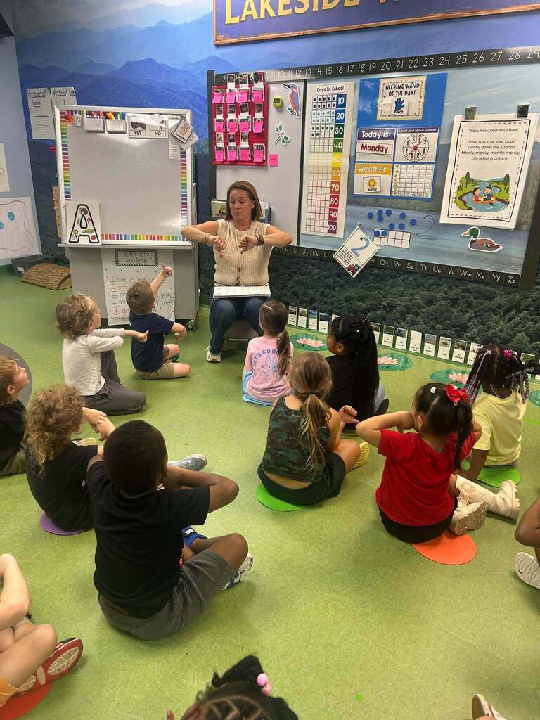 Mrs. Alexanders leads a group of young students in a phonemic awareness activity. Students sit on colorful floor spots and follow along with hand motions as the teacher models the lesson.
