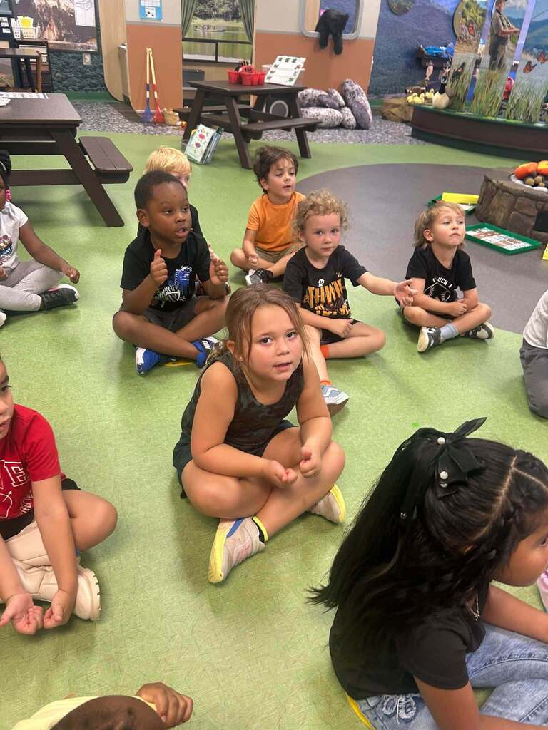 Young K4 students sit together on the classroom floor participating in a phonemic awareness lesson. Several students use hand motions and gestures as they listen and respond.