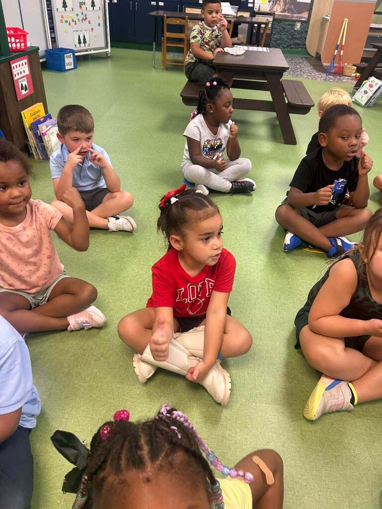 A group of young students sit together on the classroom floor, actively participating in a lesson by listening, responding, and using hand gestures.