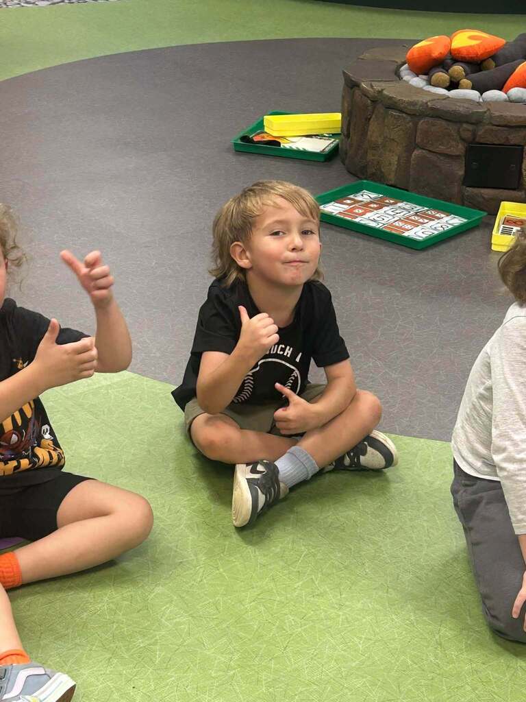 A young student sits cross-legged on the classroom floor, smiling and giving a thumbs-up while participating in a learning activity with hand motions.