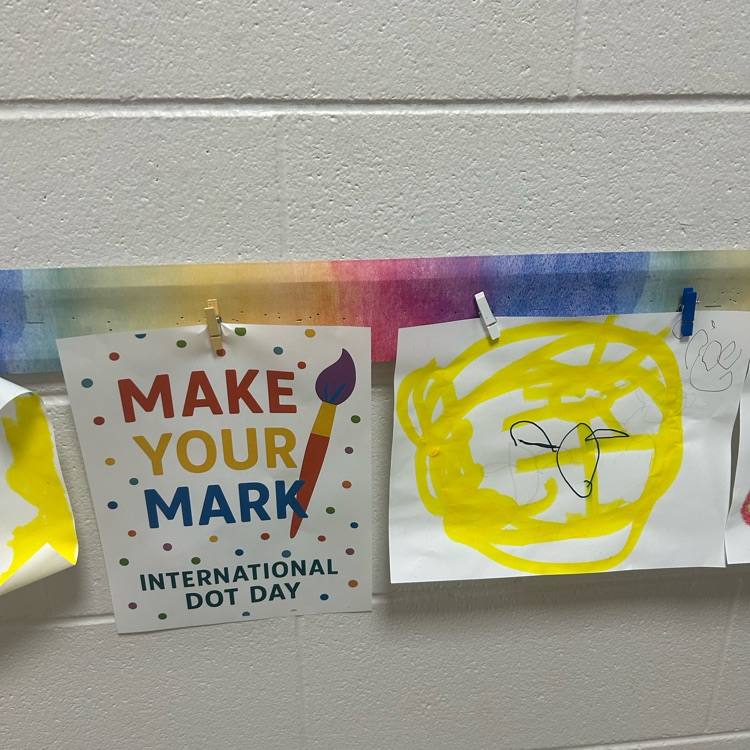 An “Inspiration Begins with a Dot” sign displayed beside a student’s artwork featuring a small black dot surrounded by a bright yellow border.