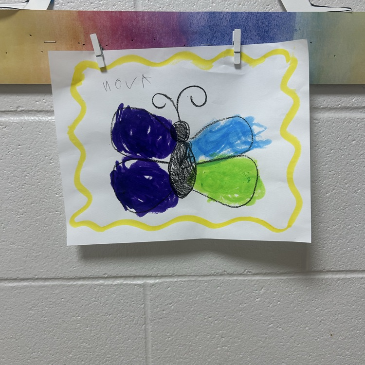 Student artwork featuring a colorful butterfly with purple, blue, and green wings, displayed with clothespins on the art room wall.