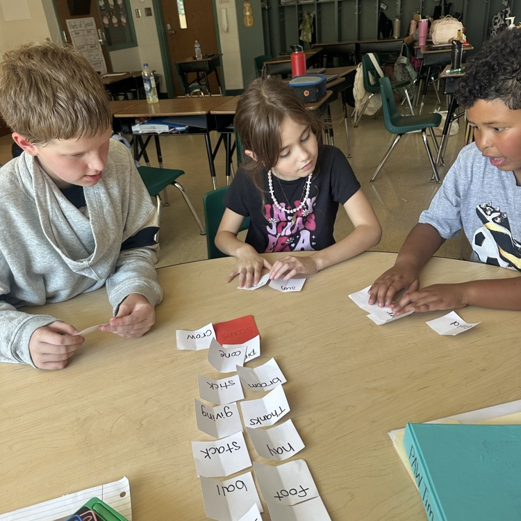 Three third grade students sit around a classroom table working on a compound word activity. Word cards are spread out on the table as the students match and sort them together during a hands-on literacy lesson in Ms. Bezy’s class.
