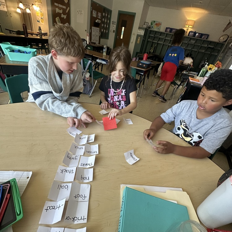Three third grade students sit around a classroom table working on a compound word activity. Word cards are spread out on the table as the students match and sort them together during a hands-on literacy lesson in Ms. Bezy’s class.
