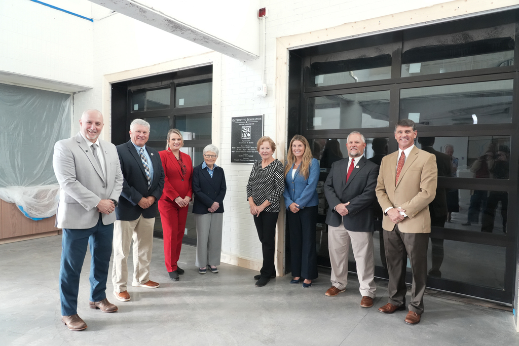 Photo of Board of Trustees members with Dr. Danny Merck, posing in front of plaque in G2i lobby