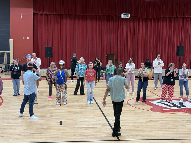 A group of adults participates in an activity on a school gymnasium floor, with several people clapping and watching. Red stage curtains, speakers, a podium, and musical instruments are visible in the background.