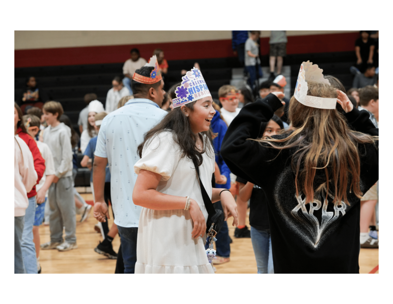 Students wearing decorated paper crowns celebrating Hispanic Heritage Month dancing and smiling during an event in a school gymnasium.