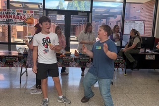Two students enjoying empanadas in front of a decorated food table during a school Hispanic Heritage Month celebration.