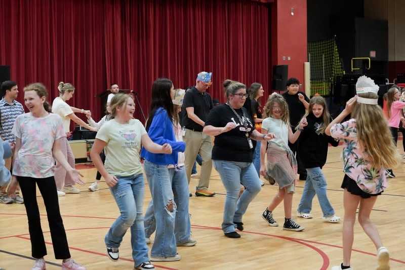 Students and staff dancing line-dance style across a wooden gymnasium floor in front of a red stage curtain. Several students wear decorated paper crowns and smile while moving to the music during a school Hispanic Heritage Month event.