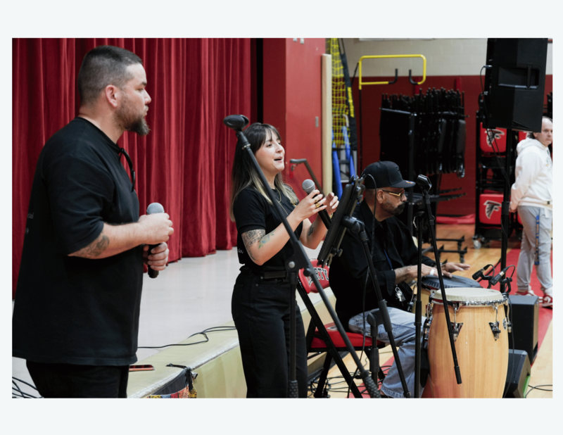 Musicians perform live with microphones and conga drums on stage during a school Hispanic Heritage Month celebration.