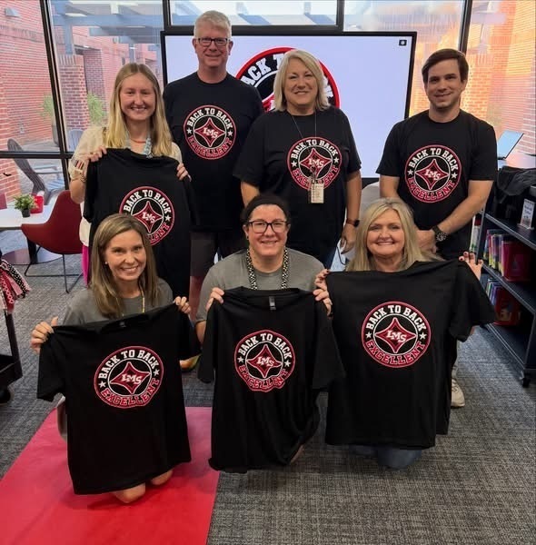Group of Liberty Middle School staff members wearing and holding black t-shirts that say "Back to Back Excellent" to celebrate their school achievement.