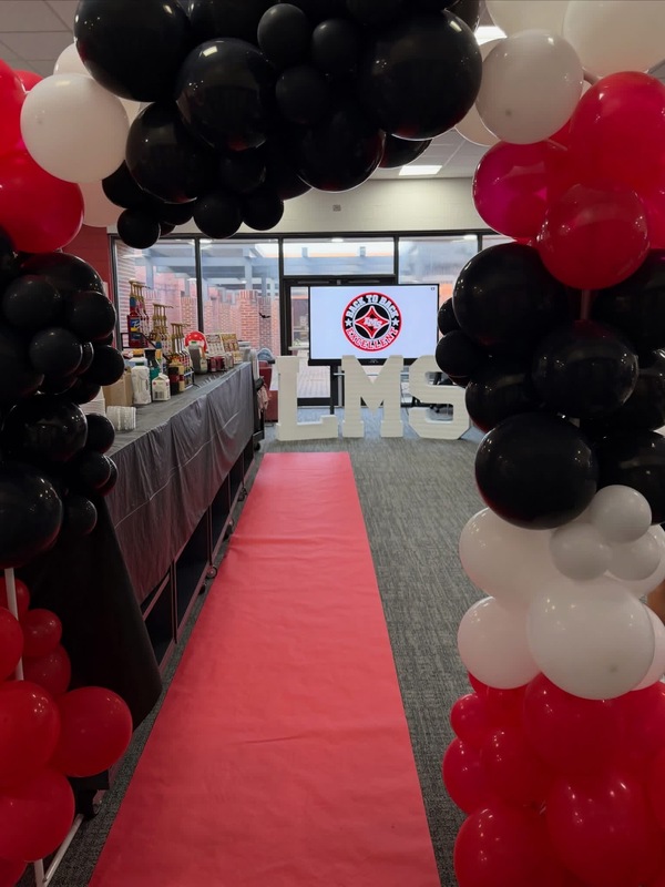 Red carpet walkway framing a celebration setup decorated with black, red, and white balloon arches, large marquee letters spelling LMS, and a beverage/snack station on the side.