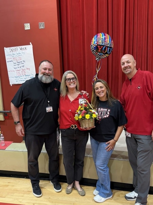 Four adults standing together in front of a stage with red curtains, holding flowers and a "Thank You" balloon.