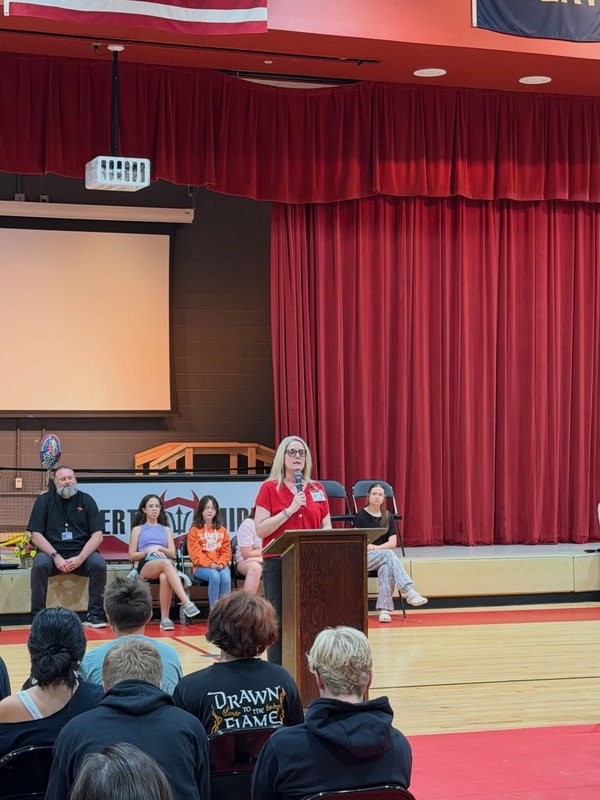 A speaker standing at a podium addressing an audience in a school gymnasium during an assembly.