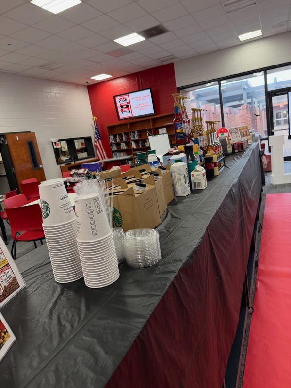 A beverage station setup in a library, featuring coffee cups, coffee travelers, flavored syrups, and creamers lined up along a long table covered with black tablecloths next to a red carpet runner.