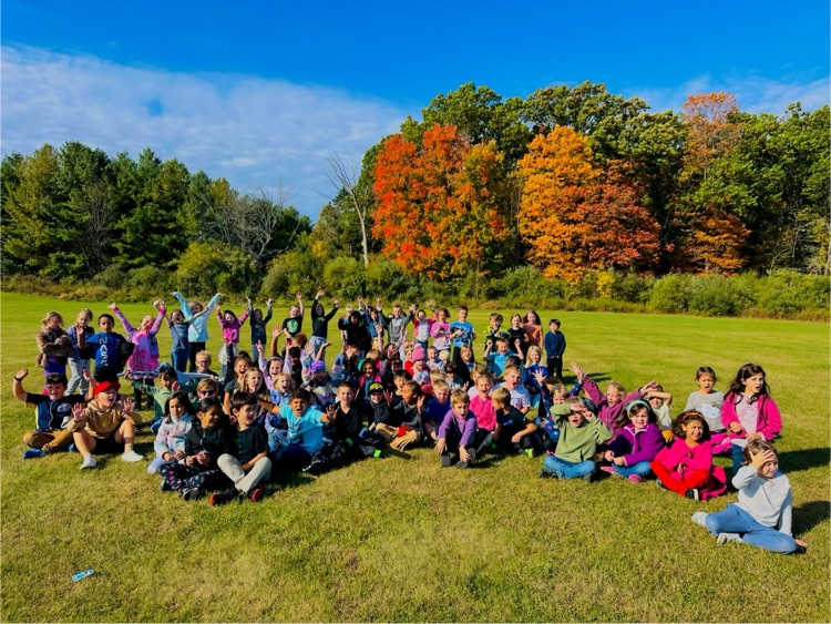 first graders sitting on the field in front of the trees. 