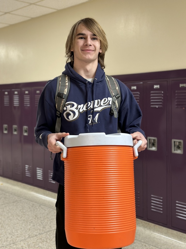 A smiling male student with long blonde hair, wearing a navy blue Milwaukee Brewers hoodie, stands in a hallway. He is holding a massive, bright orange cylindrical sports water cooler in front of him.