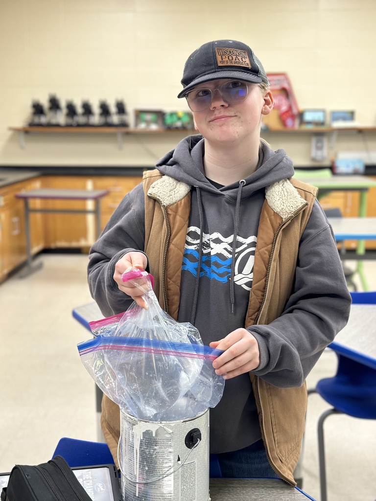 A female student wearing glasses, a cap, and a brown vest stands in a classroom. They are looking at the camera with a slight smirk while holding a metal paint can that is lined with a plastic Ziploc bag to hold their water.