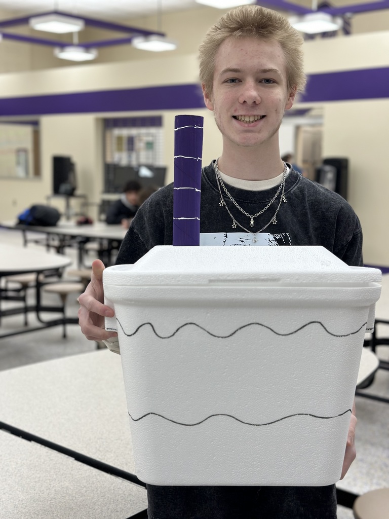 A young male student with blonde hair stands in a cafeteria smiling. He is holding a large white Styrofoam cooler that has been decorated with black wavy lines and a large purple tube sticking out of the top to resemble a giant drink cup with a straw.