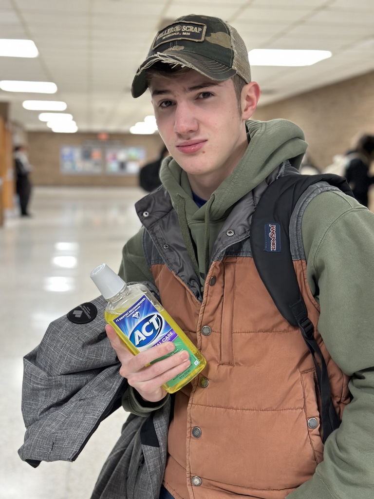 A male student wearing a camouflage baseball cap, green hoodie, and brown vest stands in a hallway. He maintains a serious expression while holding a bottle of yellow ACT mouthwash.