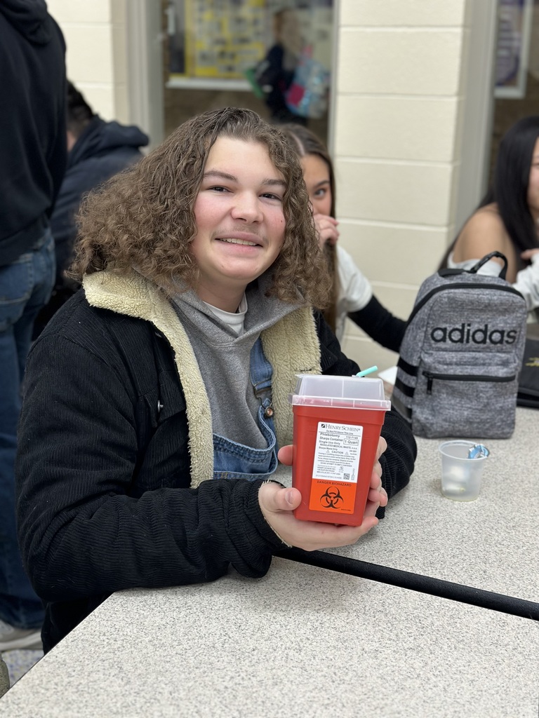 A student with shoulder-length curly hair and a denim overall outfit sits at a table smiling. They are holding a red plastic biohazard "Sharps Container" typically used for medical waste, humorously using it as a personal container.