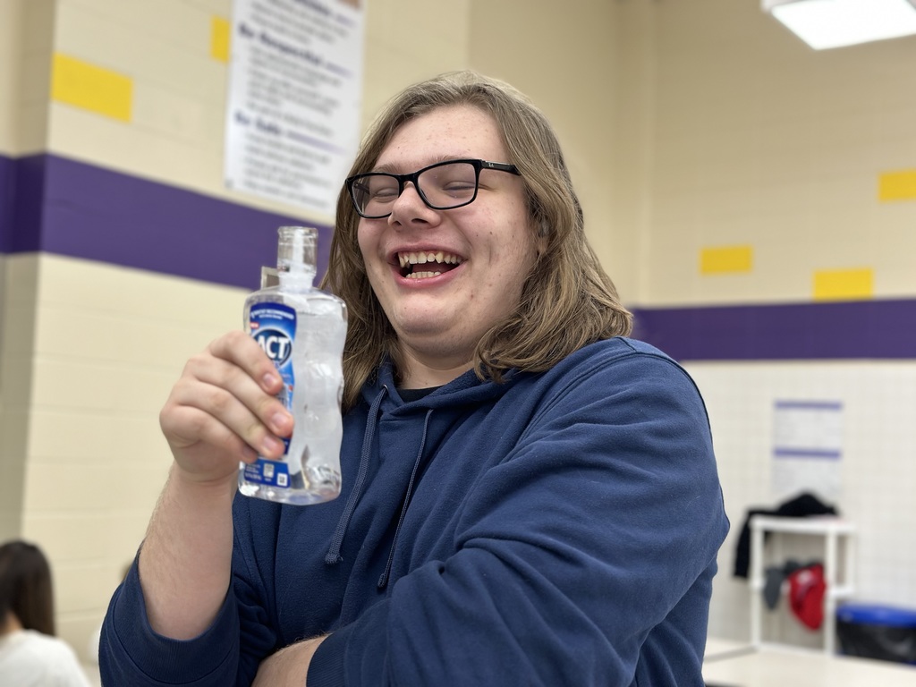 A medium shot of a laughing male student with glasses and shoulder-length hair wearing a blue hoodie. He is holding up a clear bottle of ACT mouthwash that appears to be filled with ice water, using it as a drink container in a school setting.